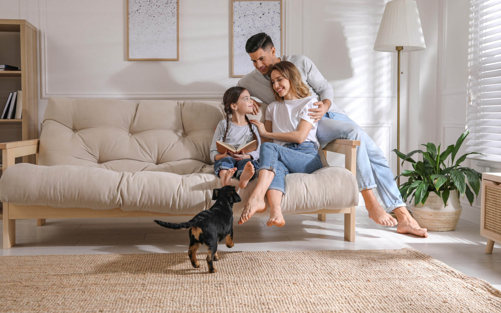 Eine junge Familie entspannt auf einem Sofa während ein kleiner Hund auf die Familie zuläuft.